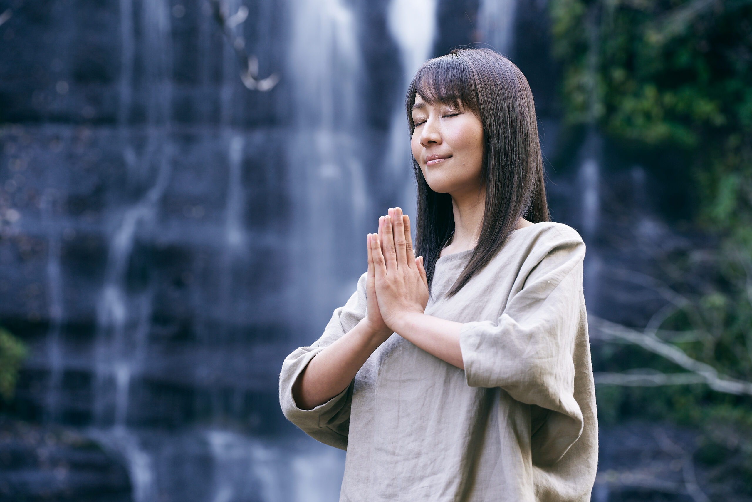滝の前で静かに合掌し、深呼吸をする女性。自然の中で心と身体がひとつになる瞬間